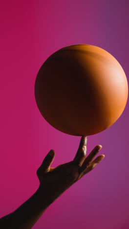 Vertical-Video-Close-Up-Studio-Shot-Of-Male-Basketball-Player-Spinning-Ball-On-Finger-Against-Pink-Lit-Background